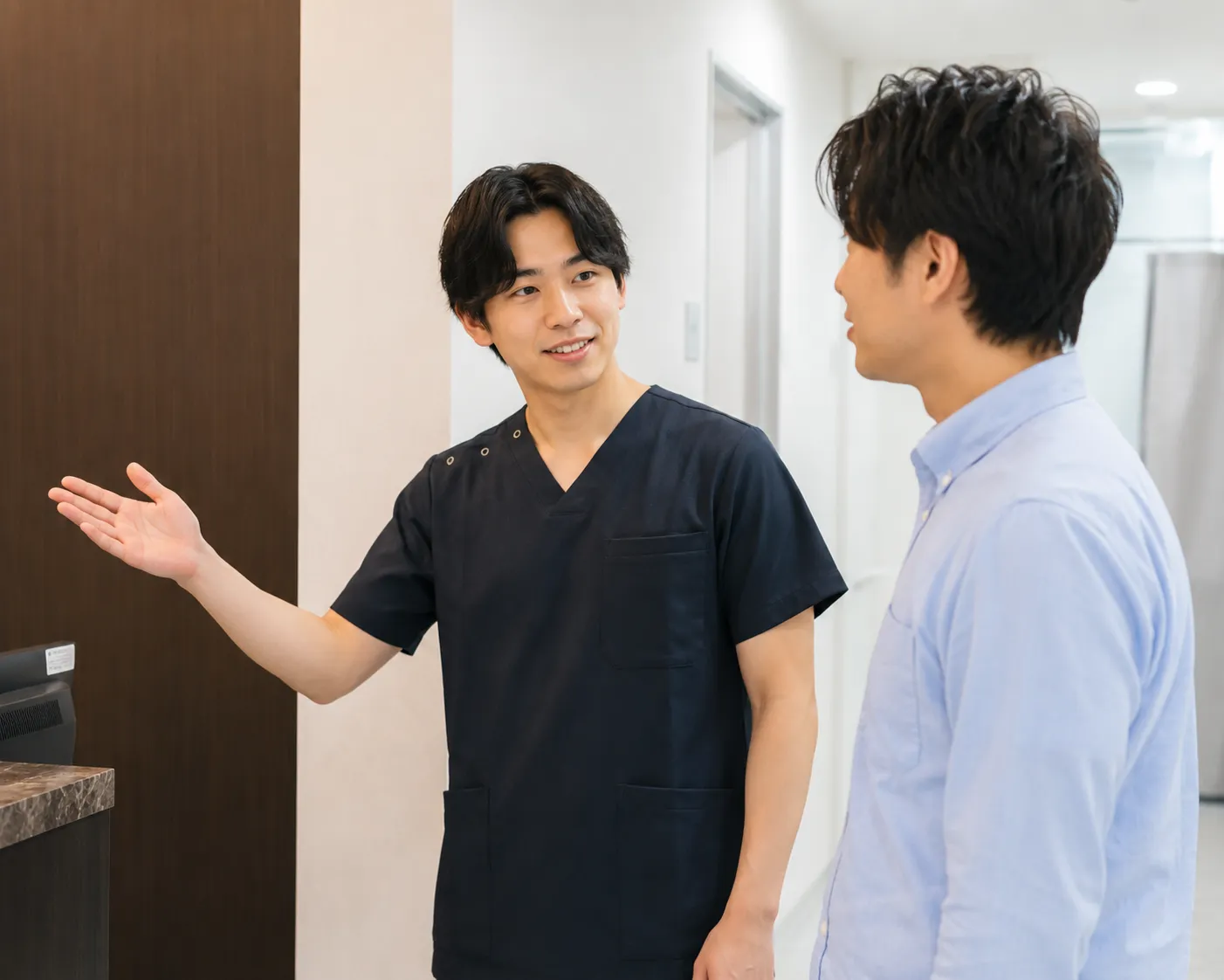 男性看護士2026年7月24日 - 美容医師・美容看護師に転職・副業なら【美医転科】(ビーテン)|医師・看護師・受付・カウンセラー Two men are chatting in a clinic hallway: a clinician in navy scrubs gestures with his left hand toward a countertop.